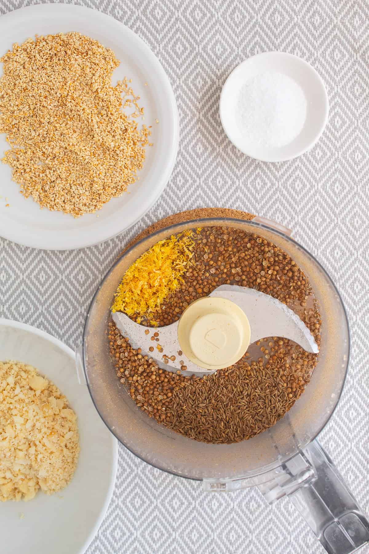 Cumin seeds, Coriander seeds, and lemon zest are added to the bowl of a food processor.
