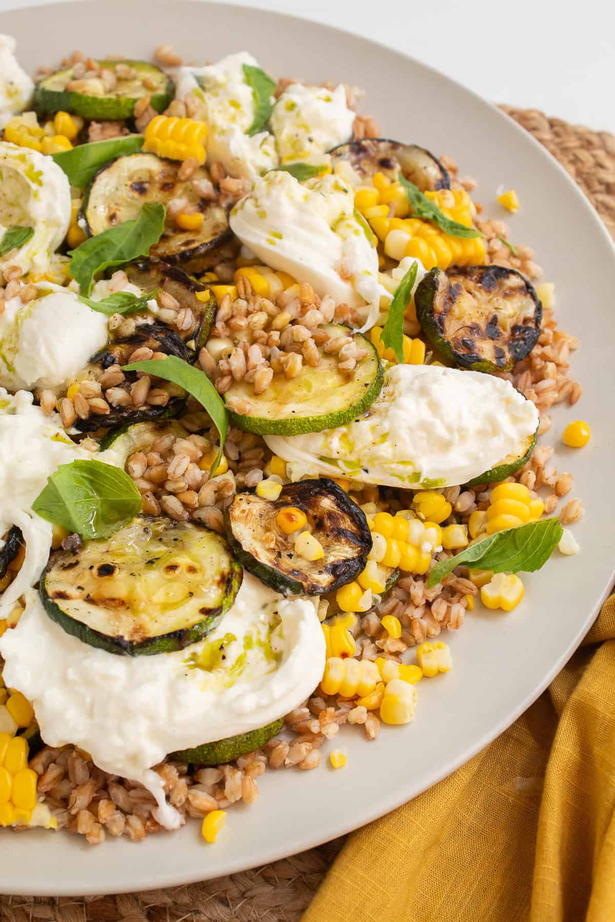The salad, with visible grains of farro, kernels of corn, grilled zucchini slices, torn basil, burrata, and green vinaigrette, is piled on a wide platter.