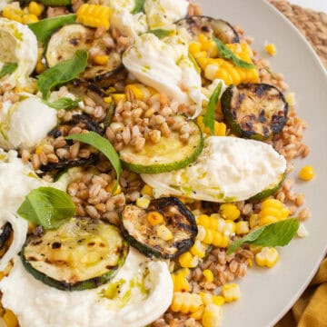 The salad, with visible grains of farro, kernels of corn, grilled zucchini slices, torn basil, burrata, and green vinaigrette, is piled on a wide platter.