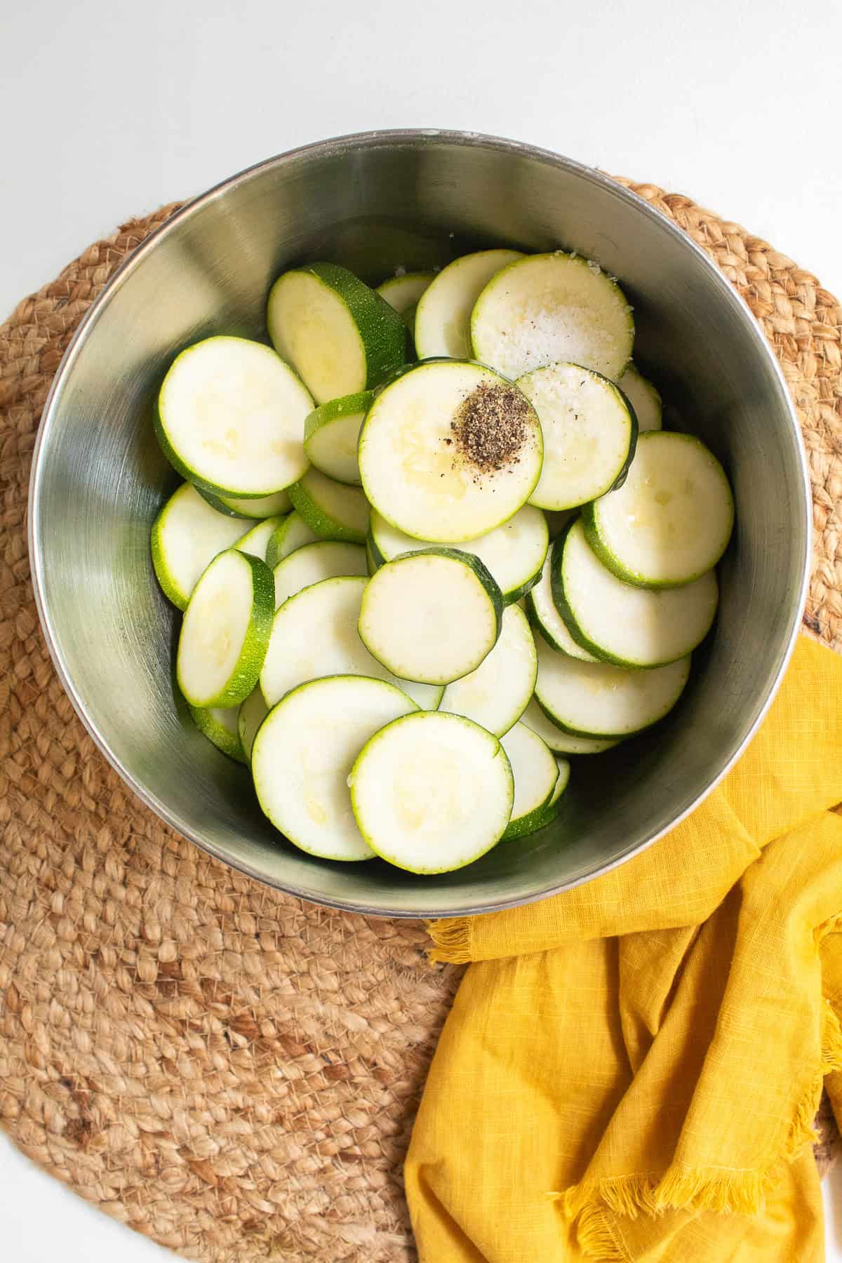 Slices of zucchini, salt, and pepper are in a stainless steel bowl.