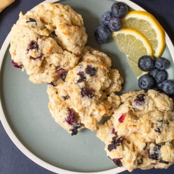 Three golden blueberry biscuits are piled on a blue plate with fresh blueberries.