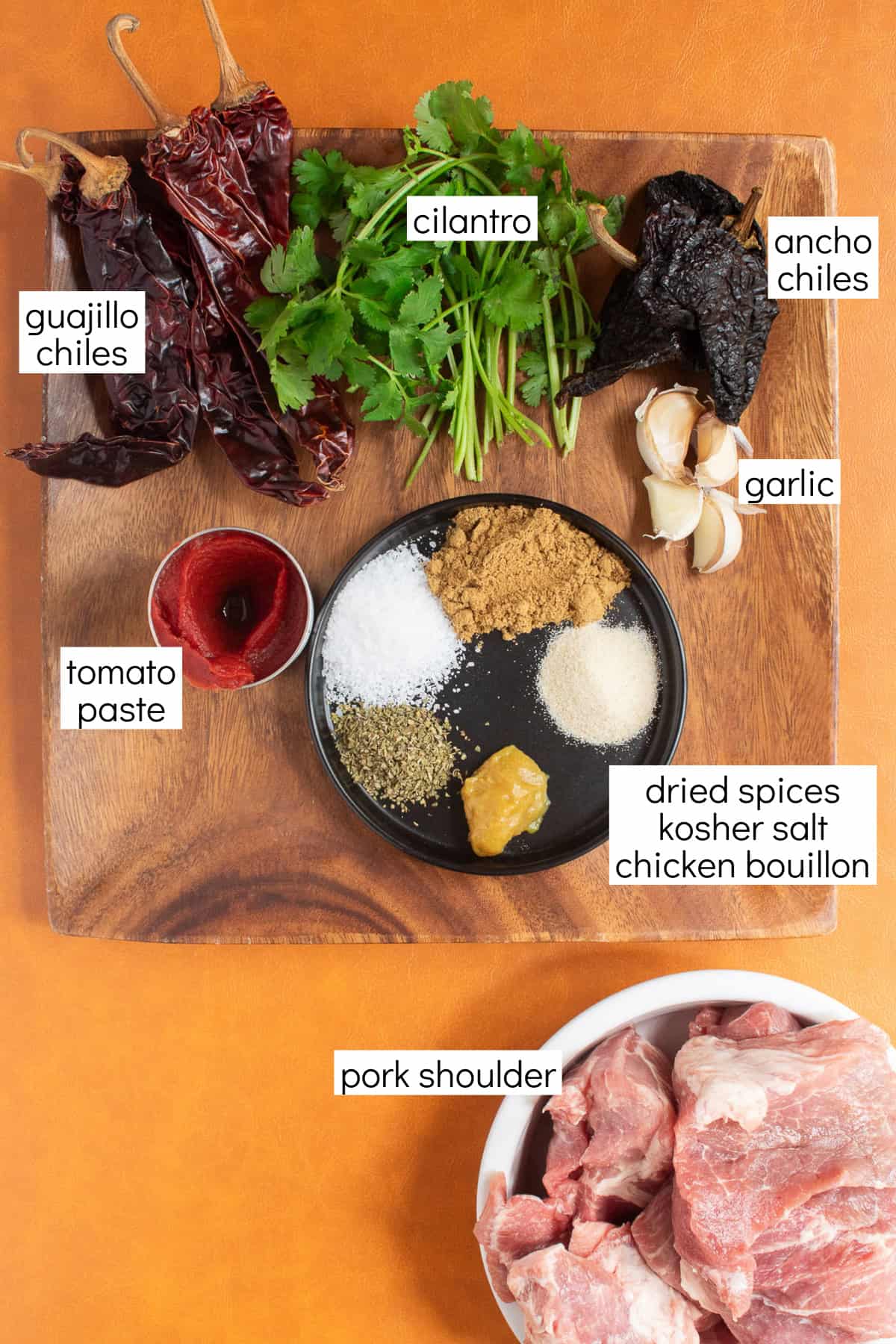 Ingredients for the recipe are displayed and labeled with white text boxes including the words guajillo chiles, cilantro, ancho chiles, tomato paste, garlic, dried spices, kosher salt, chicken bouillon, and pork shoulder.