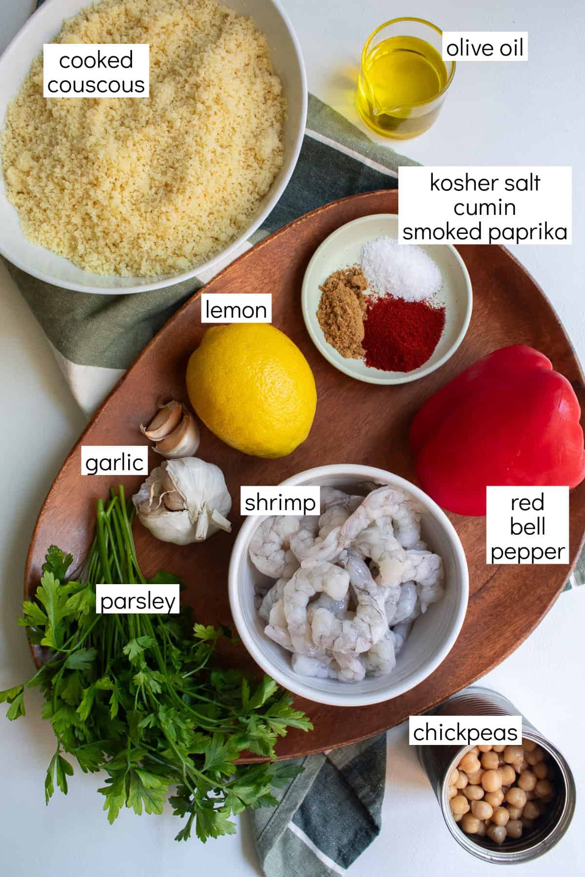 Ingredients for the recipe are displayed and labeled with white text boxes including the words cooked couscous, olive oil, kosher salt, cumin, smoked paprika, lemon, garlic, shrimp, red bell pepper, parsley, and chickpeas.