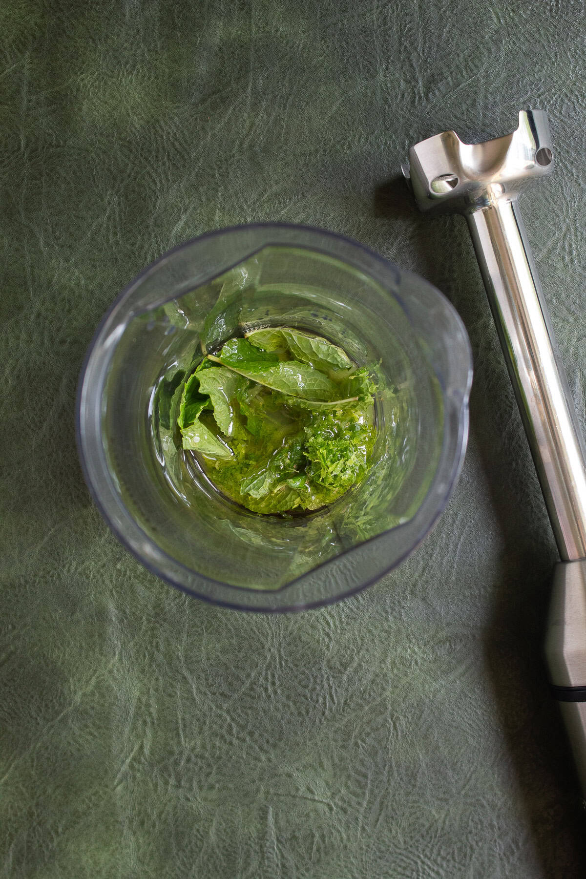 Honey, oil, mint leaves, lime juice, and lime zest are piled in an immersion blender pitcher.
