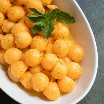 An oblong white bowl is piled with cantaloupe melon balls speckled with mint and line zest and garnished with mint leaves.