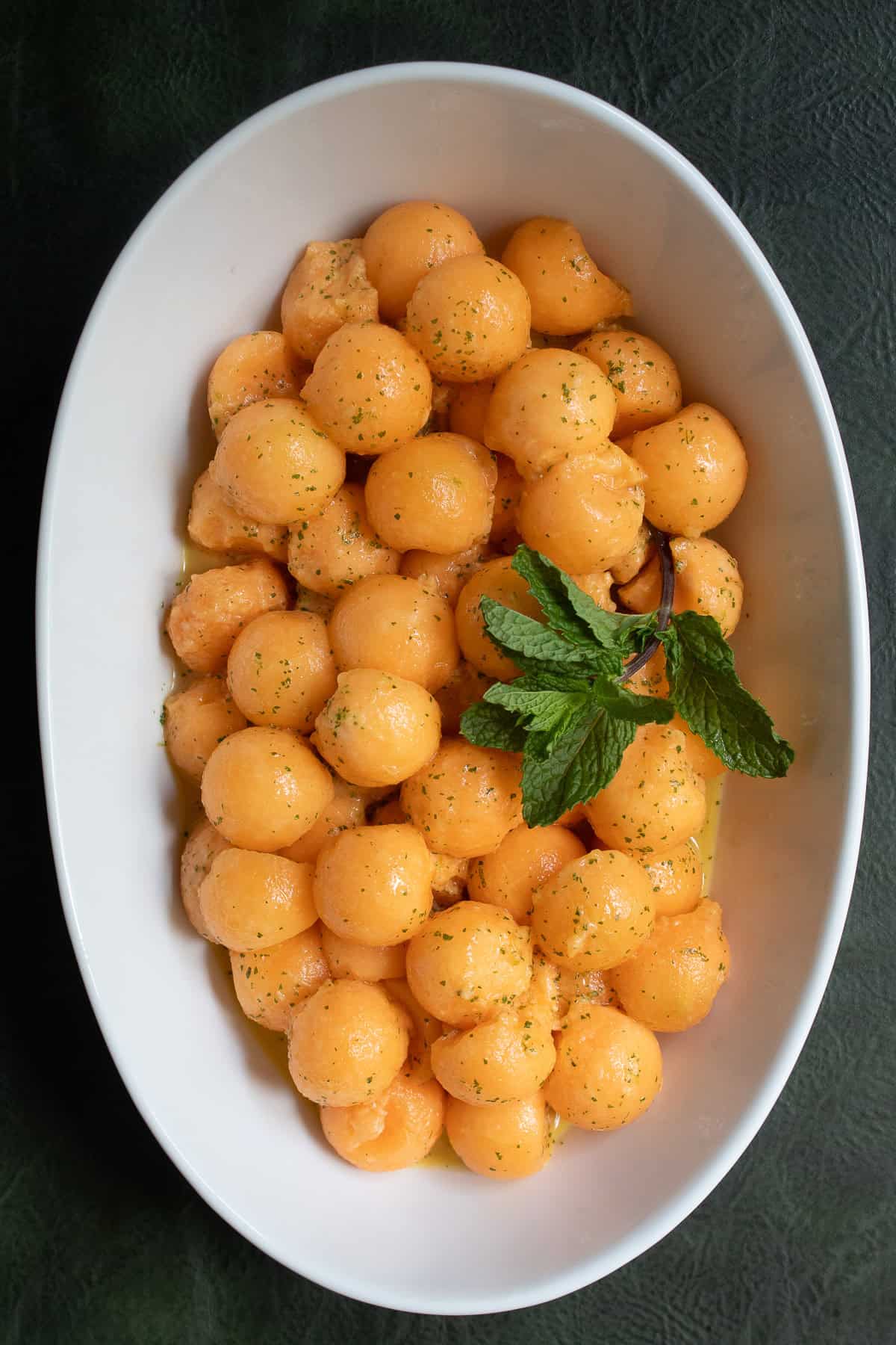 An oblong white bowl is piled with cantaloupe melon balls speckled with mint and line zest and garnished with mint leaves.