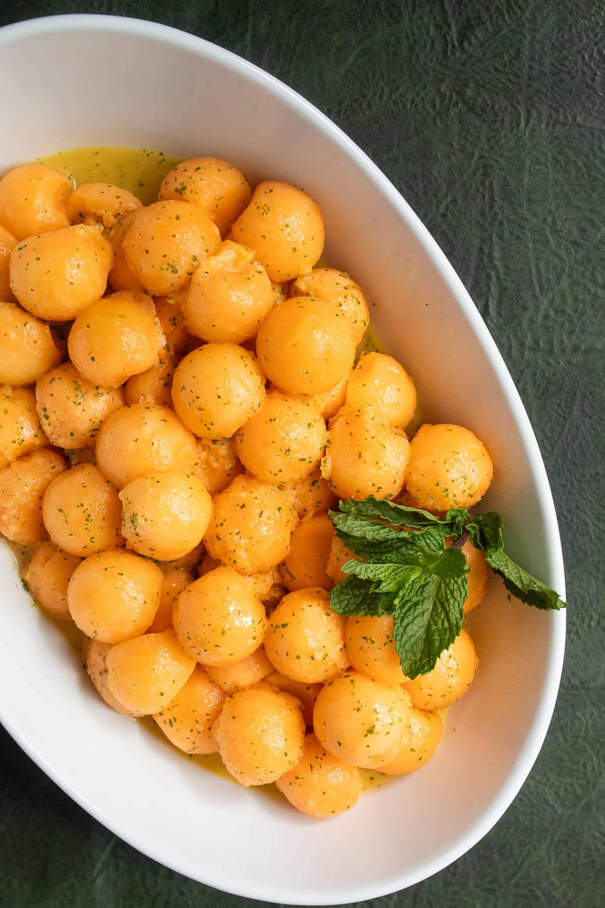 An oblong white bowl sitson a green background and is piled with cantaloupe melon balls speckled with mint and line zest and garnished with mint leaves.