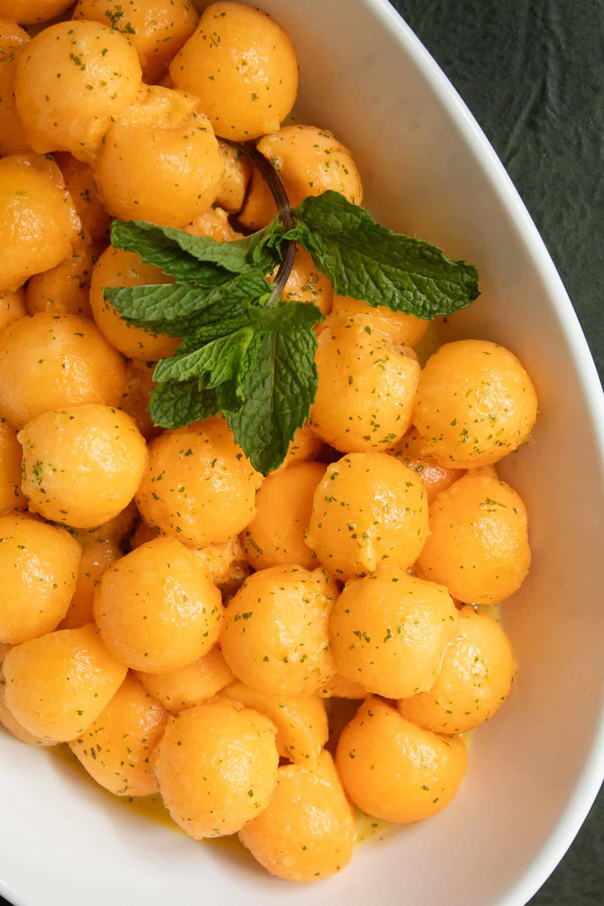 An oblong white bowl is piled with cantaloupe melon balls speckled with mint and line zest and garnished with mint leaves.