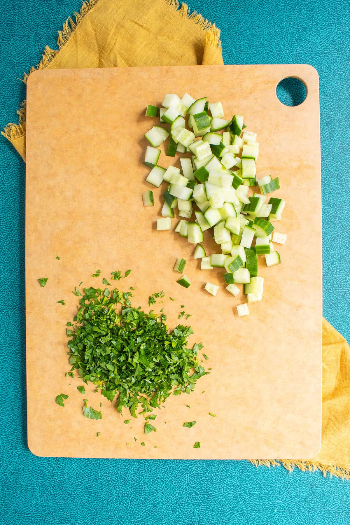 Cucumber is diced and parsley is chopped on a brown cutting board.