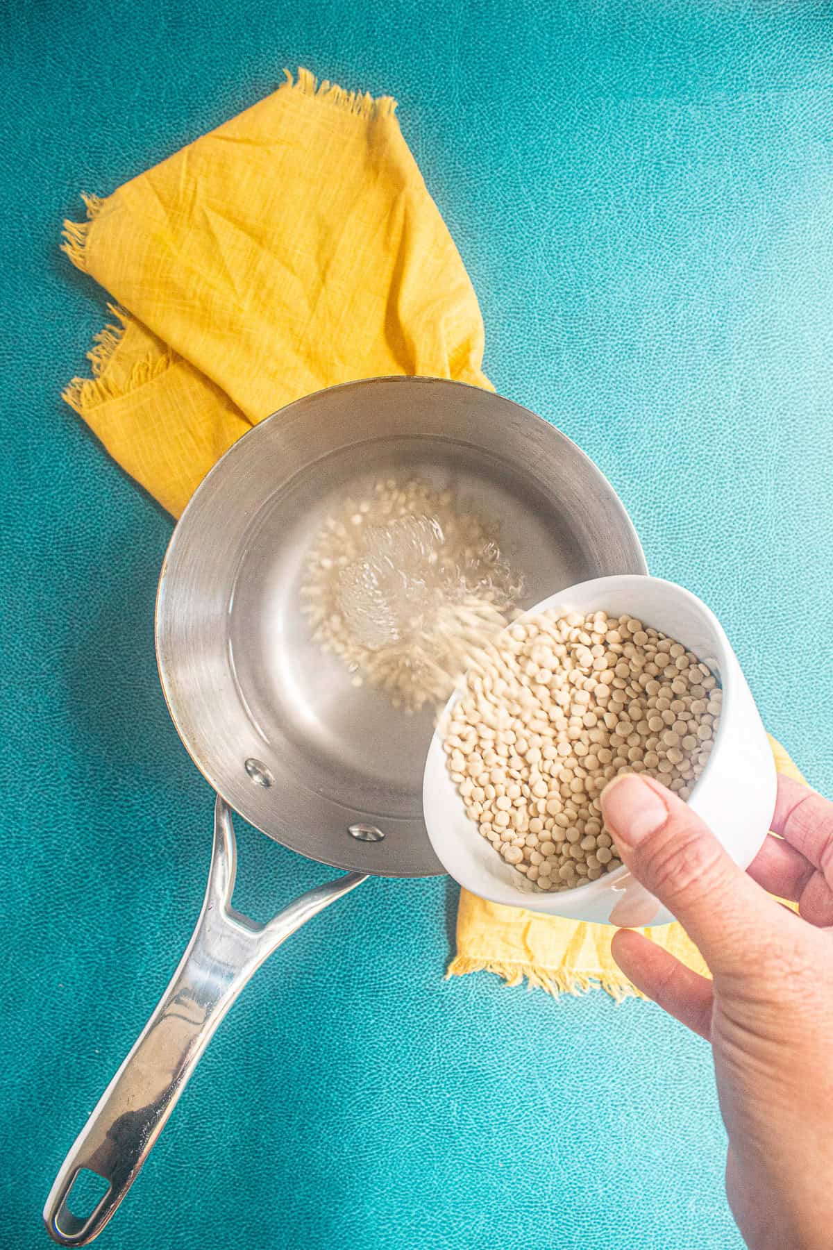 Pearl couscous is poured from a white bowl into a saucepan full of boiling water.