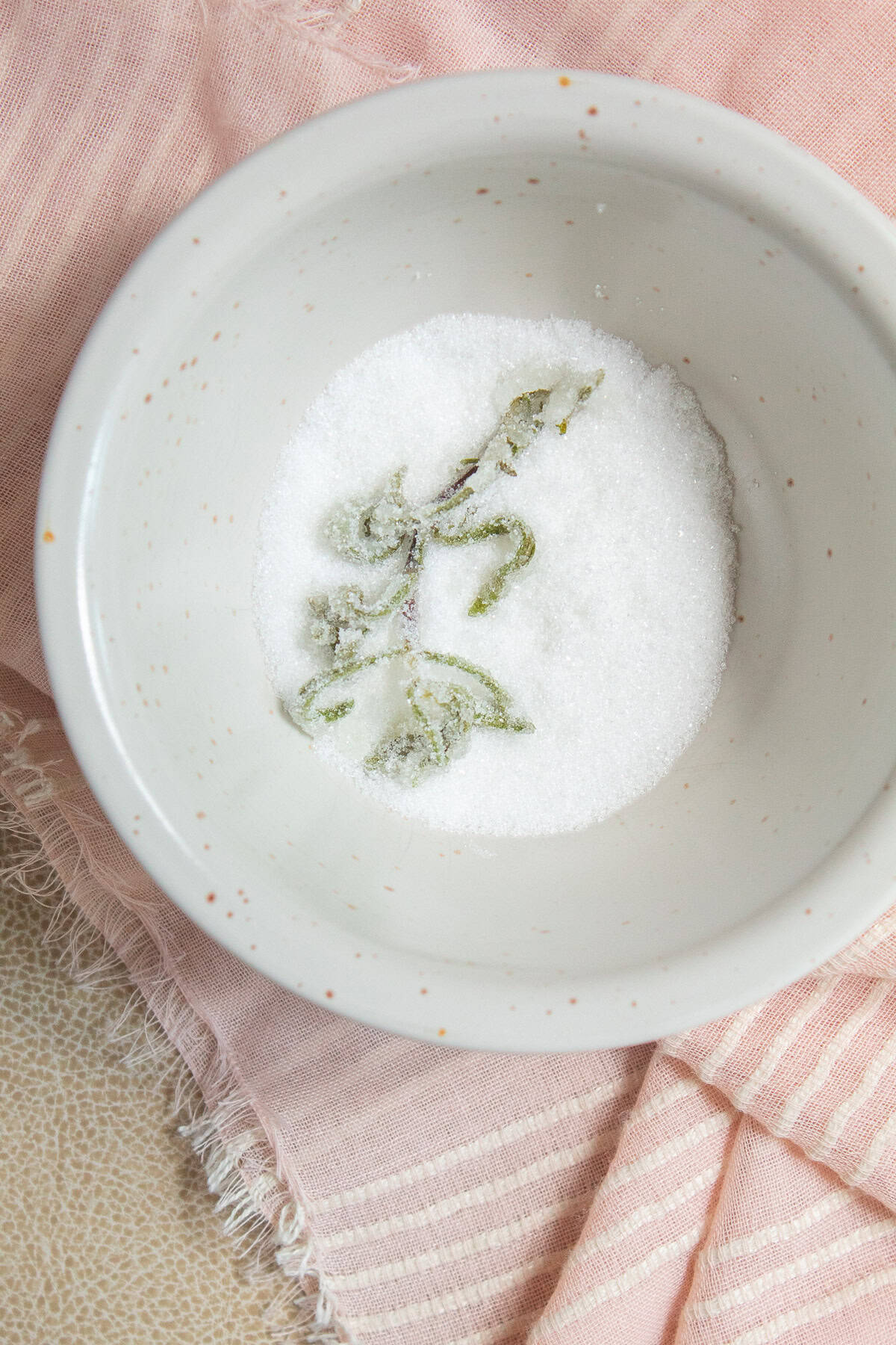 A rosemary sprig is covered with granulated sugar in a small bowl.