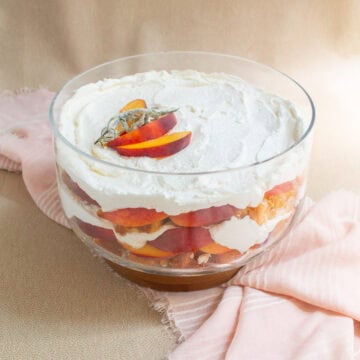 The trifle bowl is layered with angel food cake, red and yellow peaches, and whipped cream and sits on a pink and beige surface.