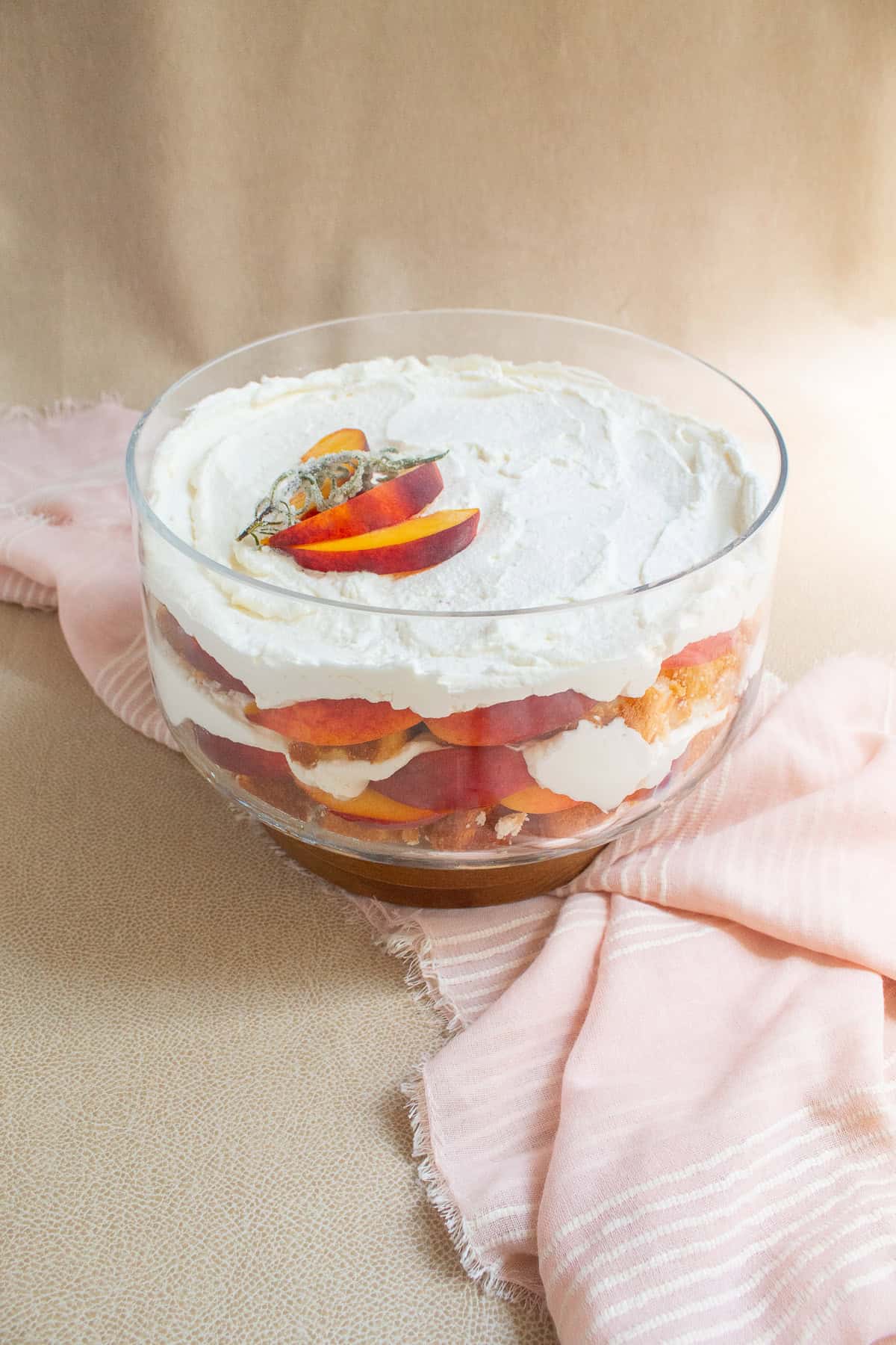 The trifle bowl is layered with angel food cake, red and yellow peaches, and whipped cream and sits on a pink and beige surface.