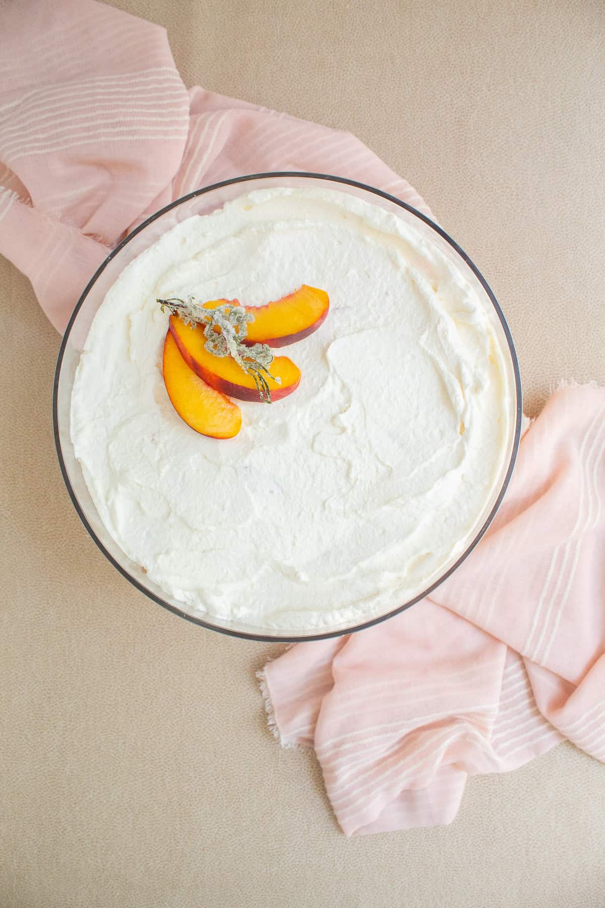 The top of the trifle is a smooth layer of whipped cream garnished with peach slices and a candied rosemary stem.