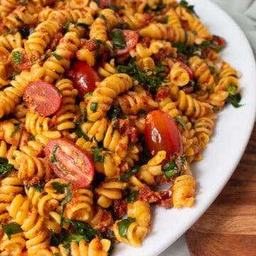 Fusilli pasta is coated with a red pesto and tossed with tomatoes, spinach, and pine nuts before being plated on a white platter.