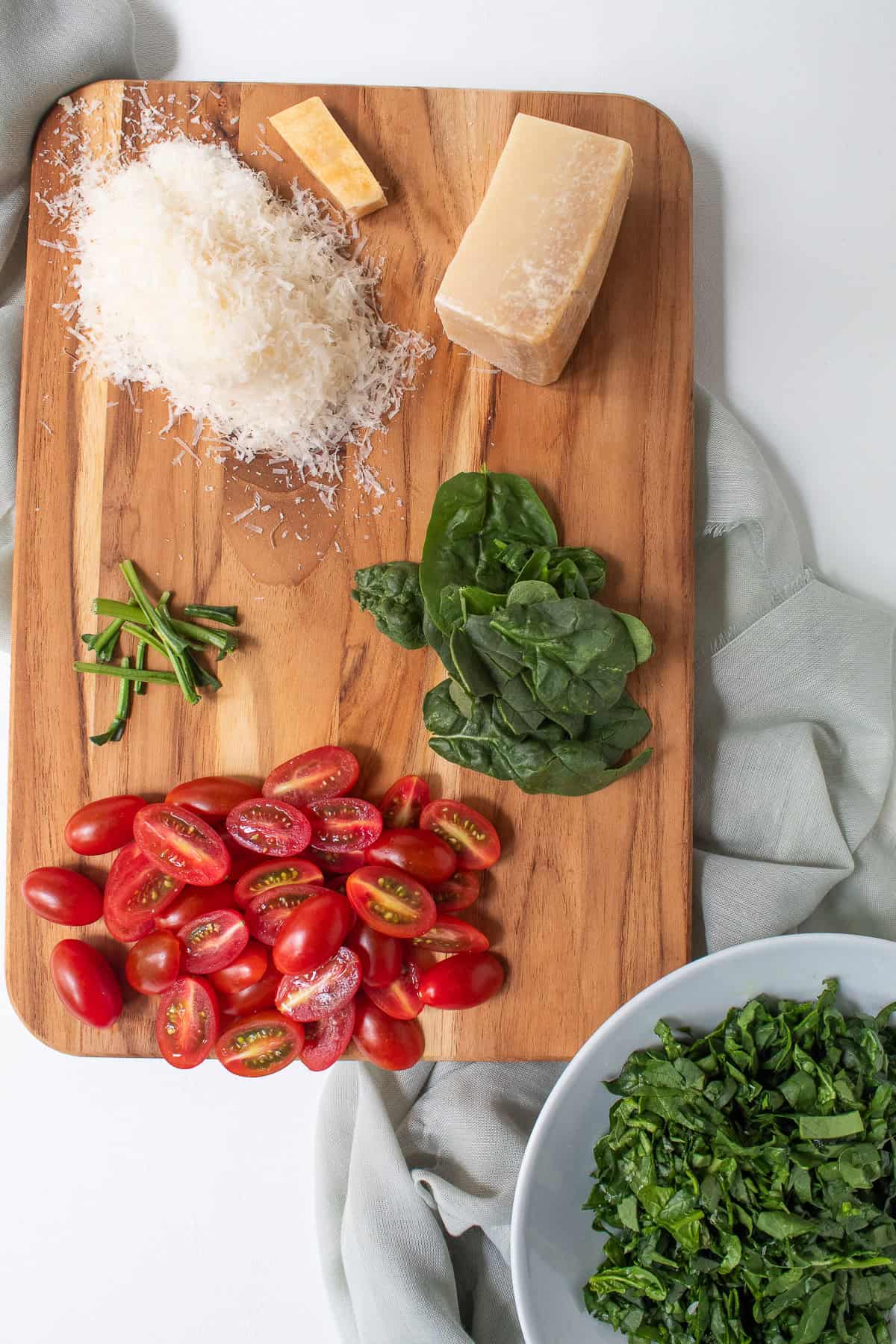 Parmesan is shredded, stems are removed from spinach leaves, and grape tomatoes are sliced in half on a wooden cutting board.