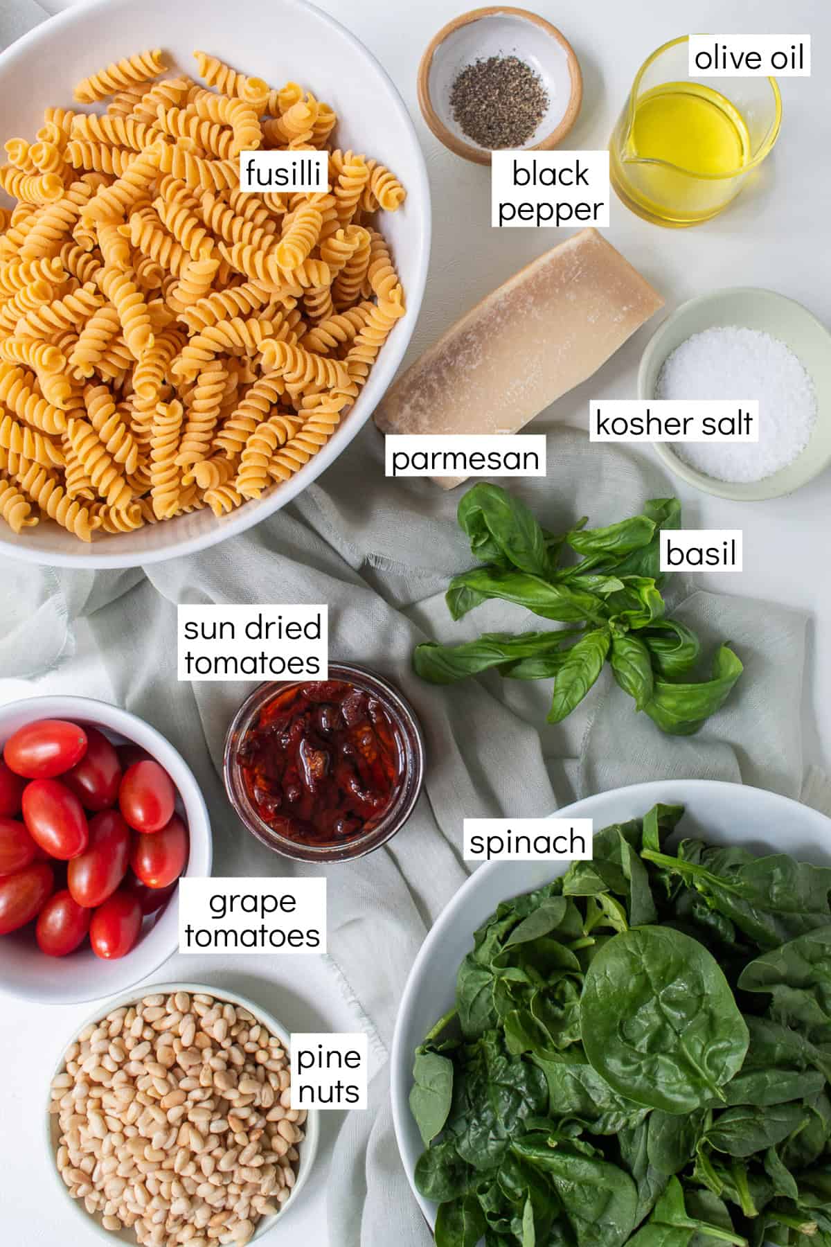 Ingredients for the recipe are displayed and labeled with white text boxes including the words fusilli, black pepper, olive oil, Parmesan, kosher salt, basil, sun dried tomatoes, grape tomatoes, spinach, and pine nuts.