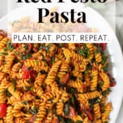 Fusilli pasta coated with a red pesto sauce is tossed with spinach, sun dried tomatoes, and fresh tomatoes and piled onto a white platter. A white text box at the top of the image contains the words, "red pesto pasta" and "plan. eat. post. repeat."