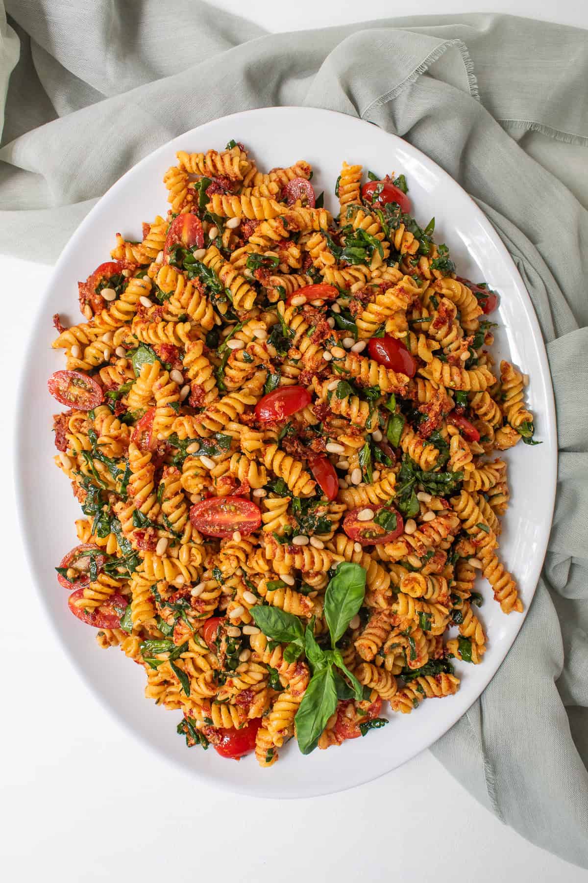A large white oval platter is piled high with the red pesto pasta and garnished with basil leaves.