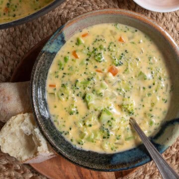 A bowl of creamy broccoli cheddar soup studded with visible broccoli and carrots sits in a gray and blue bowl next to pieces of bread.