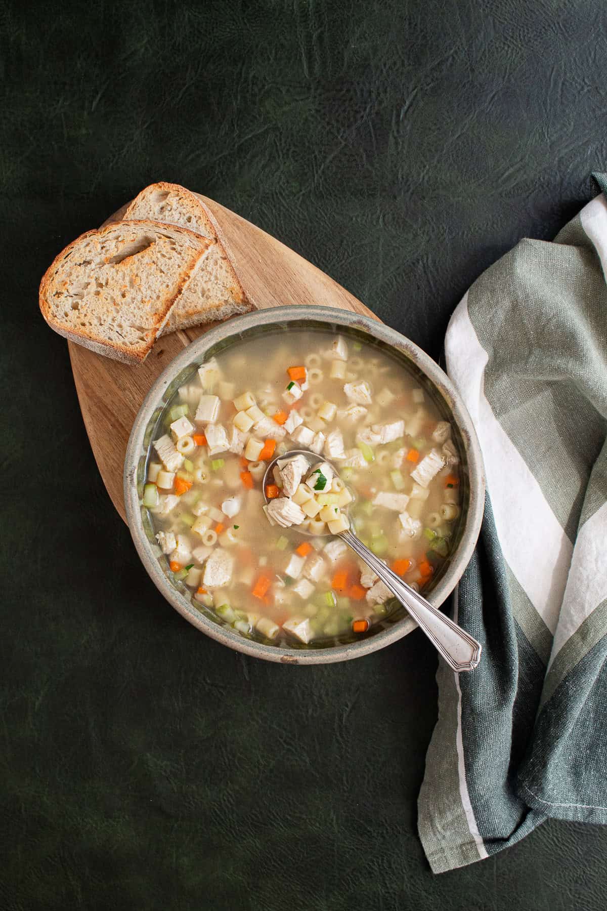 A bowl of soup with ditalini noodles, chicken chunks, carrots, and celery is served with toasted bread on a green background.