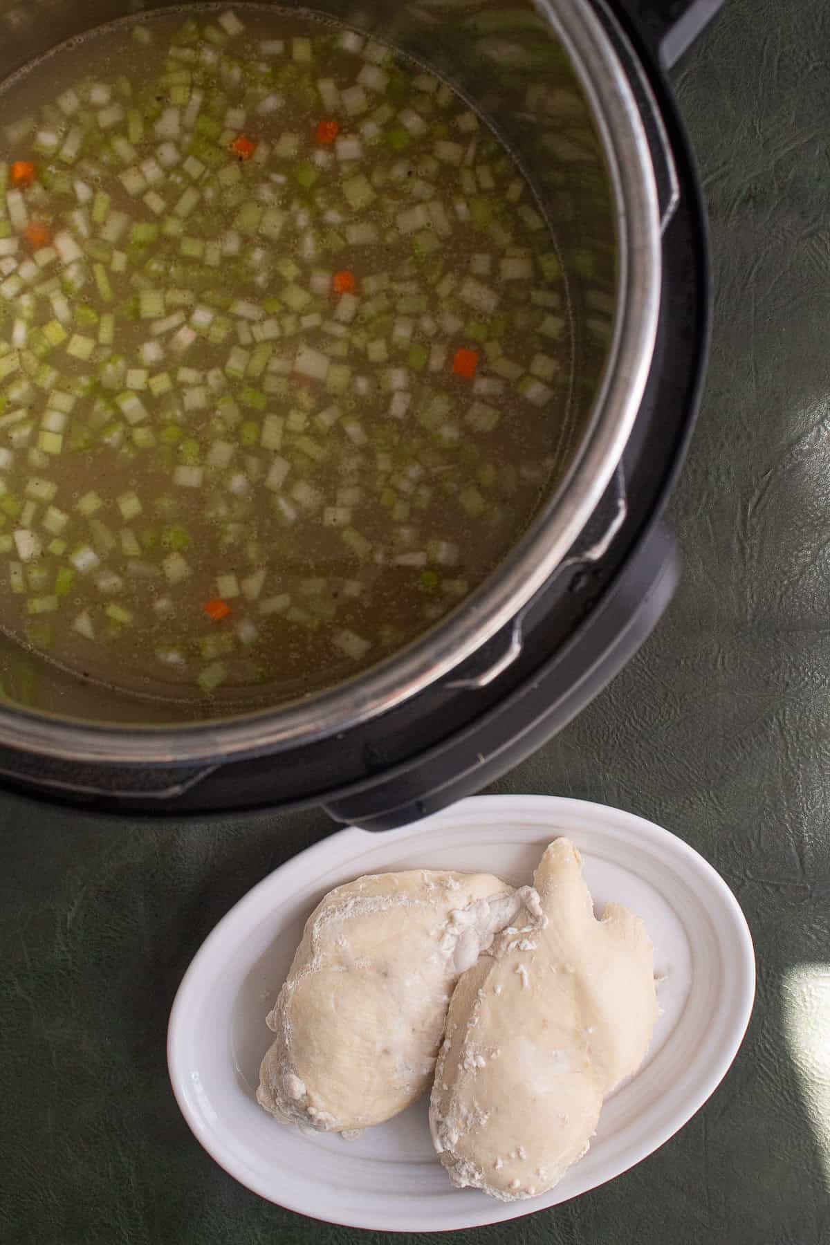 The poached chicken is on a white plate next to the pressure cooker with the pasta and vegetables.