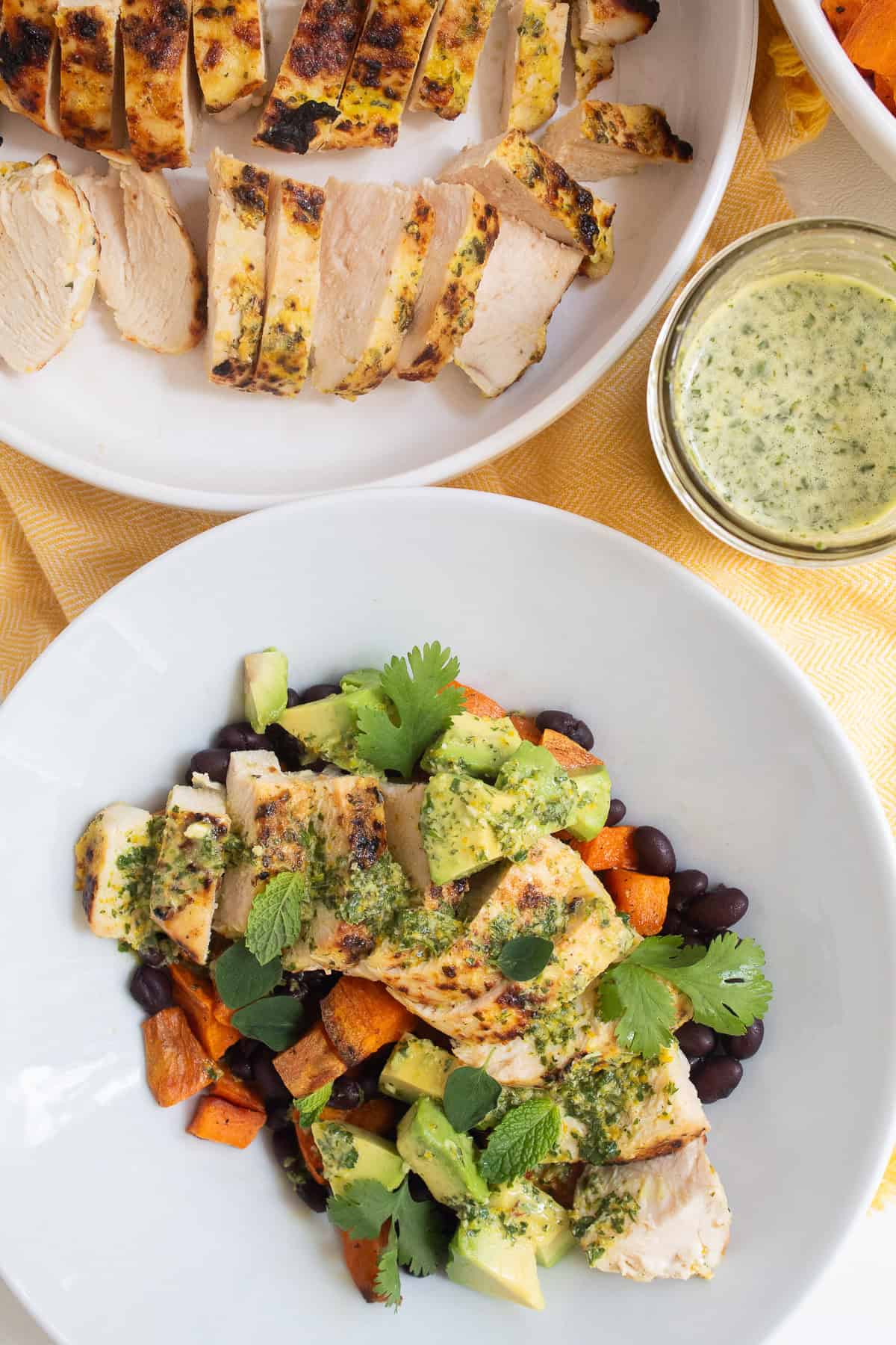 A white bowl is piled high with black beans, roasted sweet potato, mojo chicken, avocado, herbs, and mojo sauce and sits next to extra sauce in a jar.