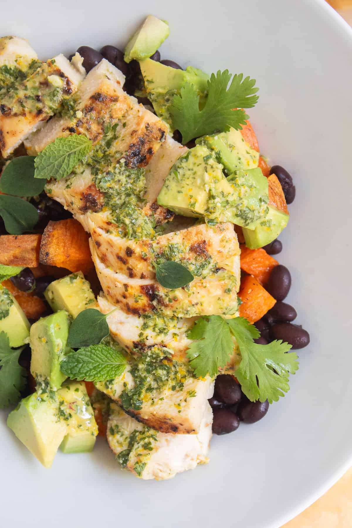 A white bowl is piled high with black beans, roasted sweet potato, mojo chicken, avocado, herbs, and mojo sauce.