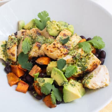 A white bowl is piled high with black beans, roasted sweet potato, mojo marinated chicken, avocado, herbs, and sauce.