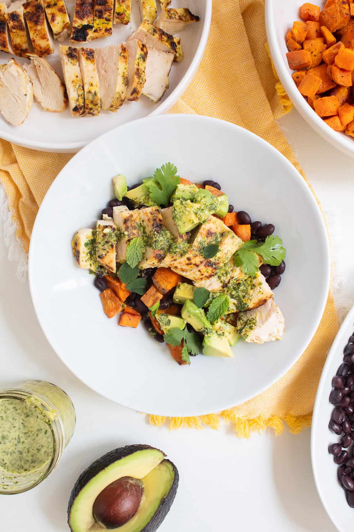 A white bowl is piled high with black beans, roasted sweet potato, mojo marinated chicken, avocado, herbs, and sauce.