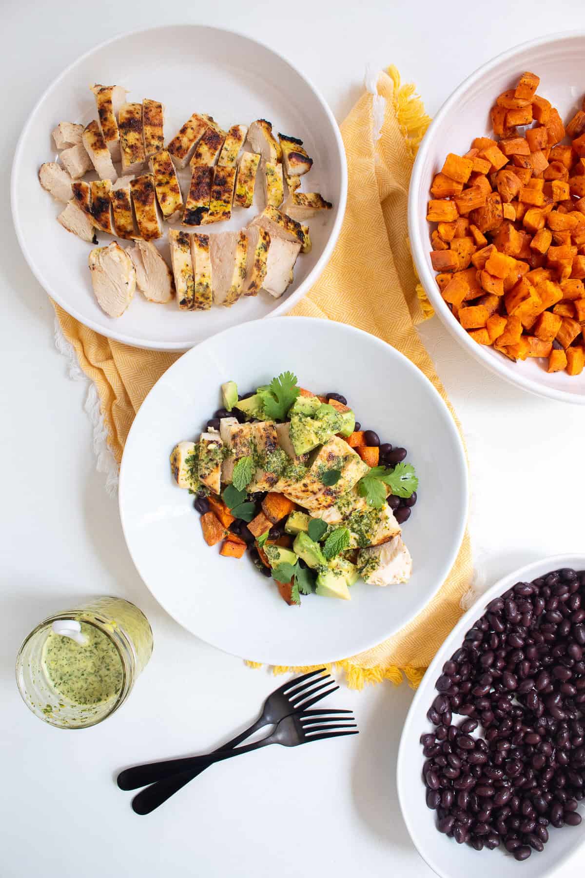 A white bowl is piled high with black beans, roasted sweet potato, mojo marinated chicken, avocado, herbs, and sauce.