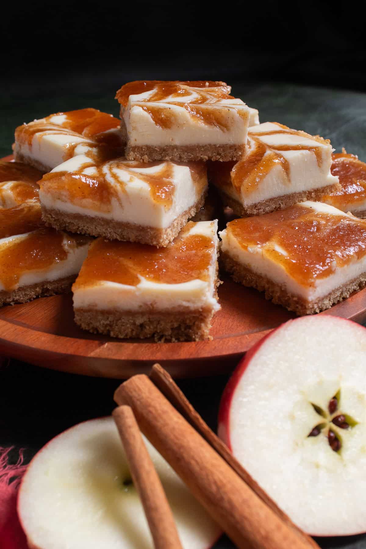Apple cheesecake bars are piled on a wooden platter next to apple slices and cinnamon sticks.