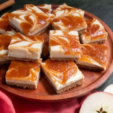 Apple cheesecake bars are piled on a wooden platter next to apple slices and cinnamon sticks.