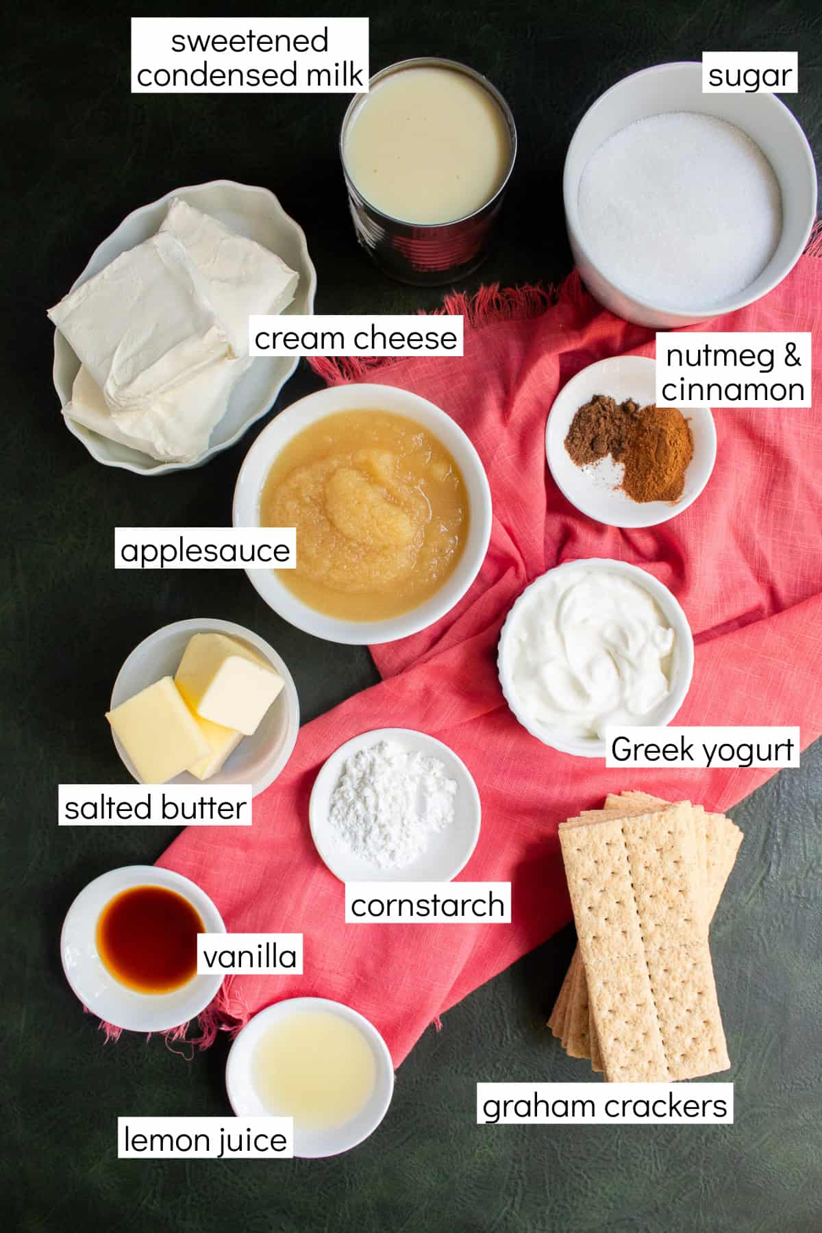 Ingredients for the recipe are displayed and labeled with white text boxes including the words sweetened condensed milk, sugar, cream cheese, nutmeg & cinnamon, applesauce, Greek yogurt, salted butter, cornstarch, vanilla, lemon juice, and graham crackers.