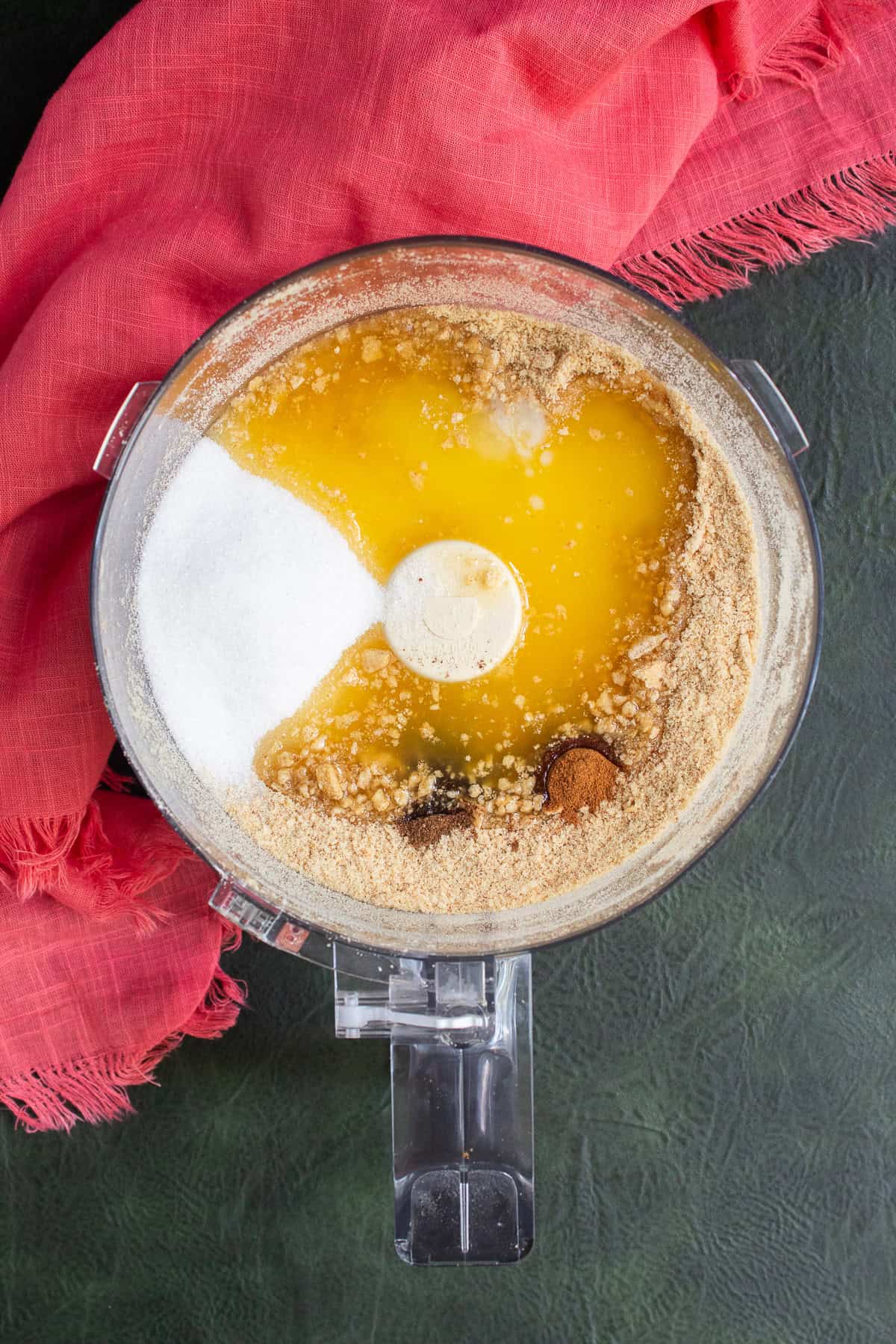 Butter, sugar nutmeg, and cinnamon are added to the graham cracker crumbs in the bowl of a food processor.