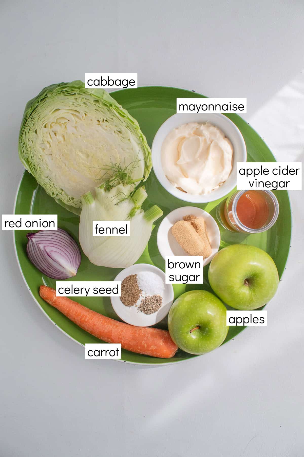 Ingredients for the recipe are displayed and labeled with white text boxes including the words cabbage, mayonnaise, apple cider vinegar, red onion, fennel, brown sugar, apples, celery seed, and carrot.