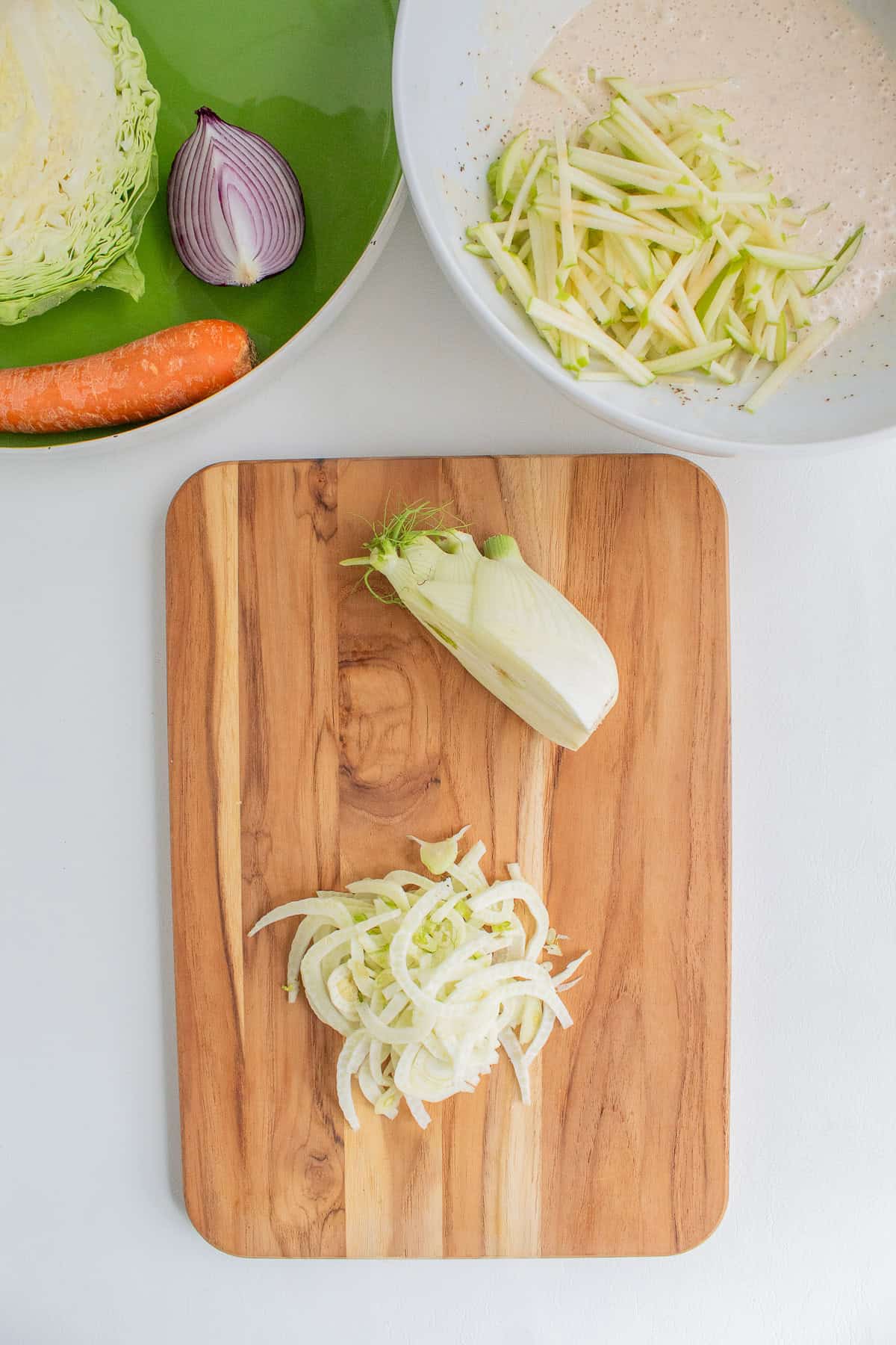 A bulb of fennel is sliced into thin strips on a wooden cutting board.