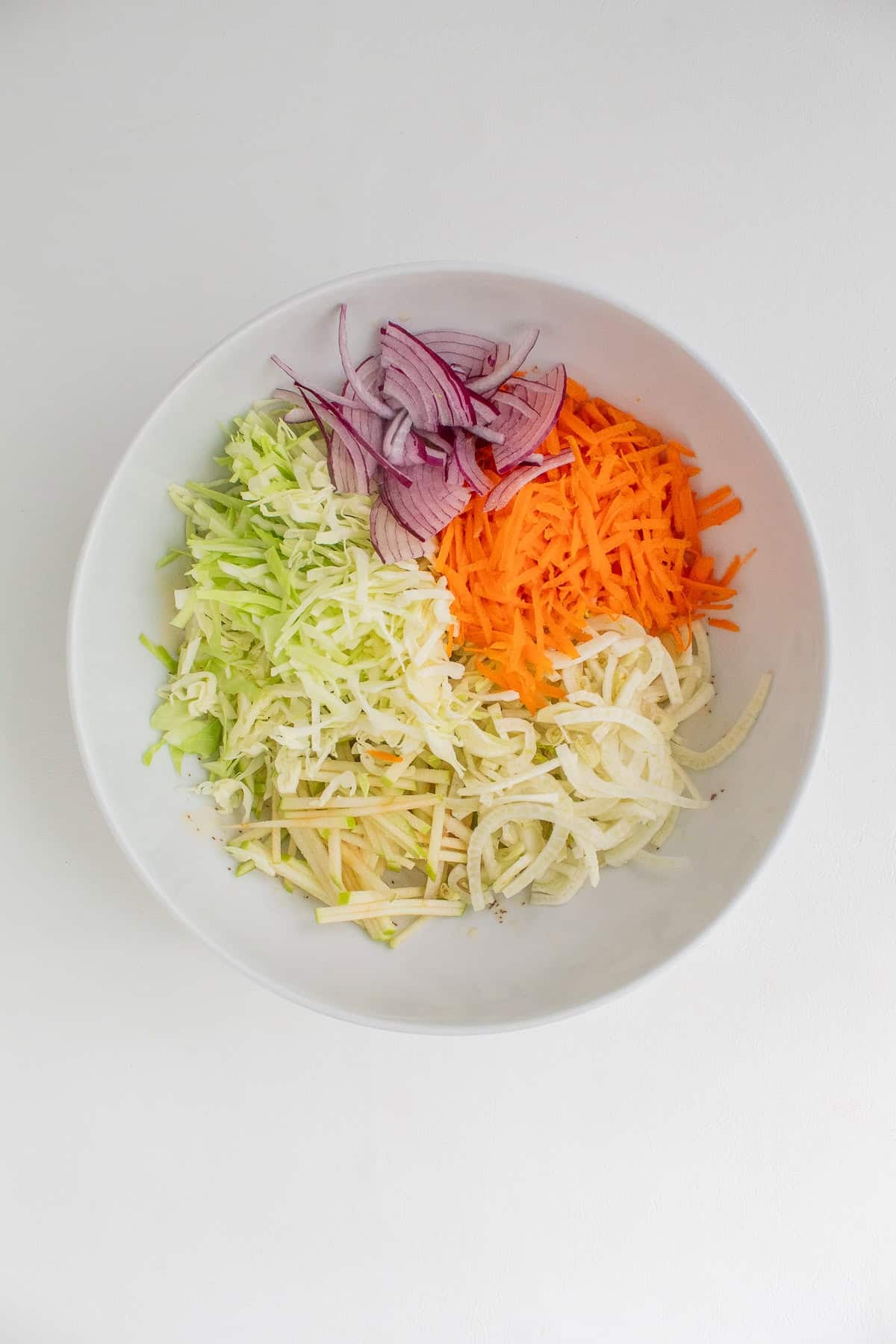 All of the shredded vegetables are plied on top of the dressing in a white bowl.