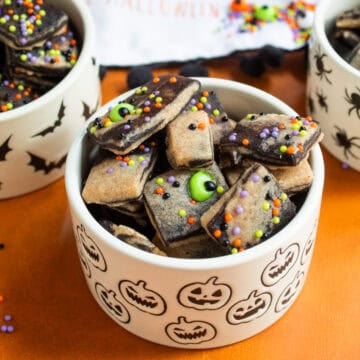Small marbled shortbread cookies decorated with sprinkles and candy eyeballs are piled into a white bowl printed with pumpkins.