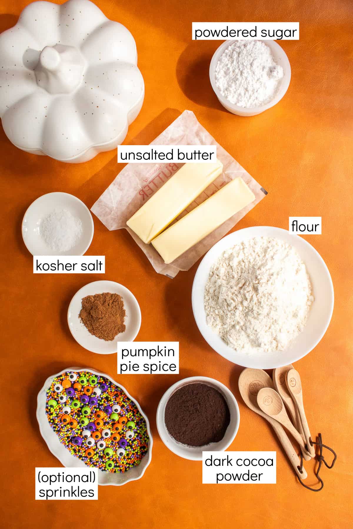 Ingredients for the recipe are displayed and labeled with white text boxes including the words powdered sugar, unsalted butter, kosher salt, flour, pumpkin pie spice, (optional) sprinkles, and dark cocoa powder.