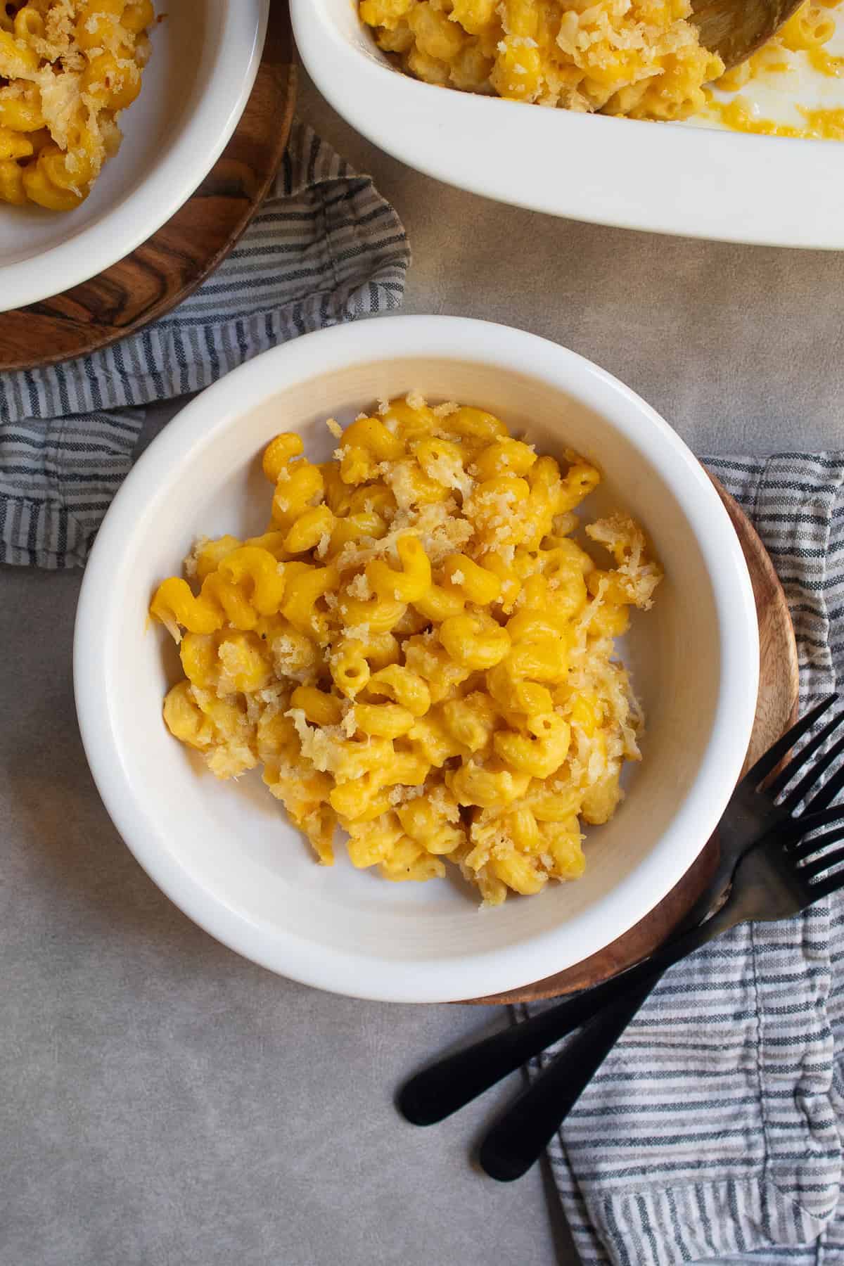 A serving of golden macaroni pasta with breadcrumb topping sits in a low white bowl next to two black forks.