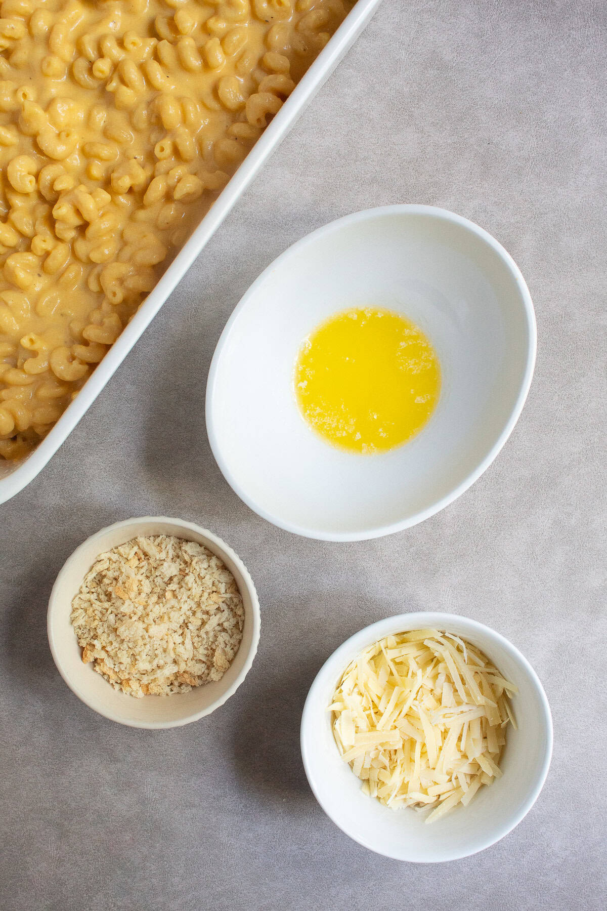 Melted butter, panko breadcrumbs, and shredded cheese sit in separate bowls on a stone-colored surface.