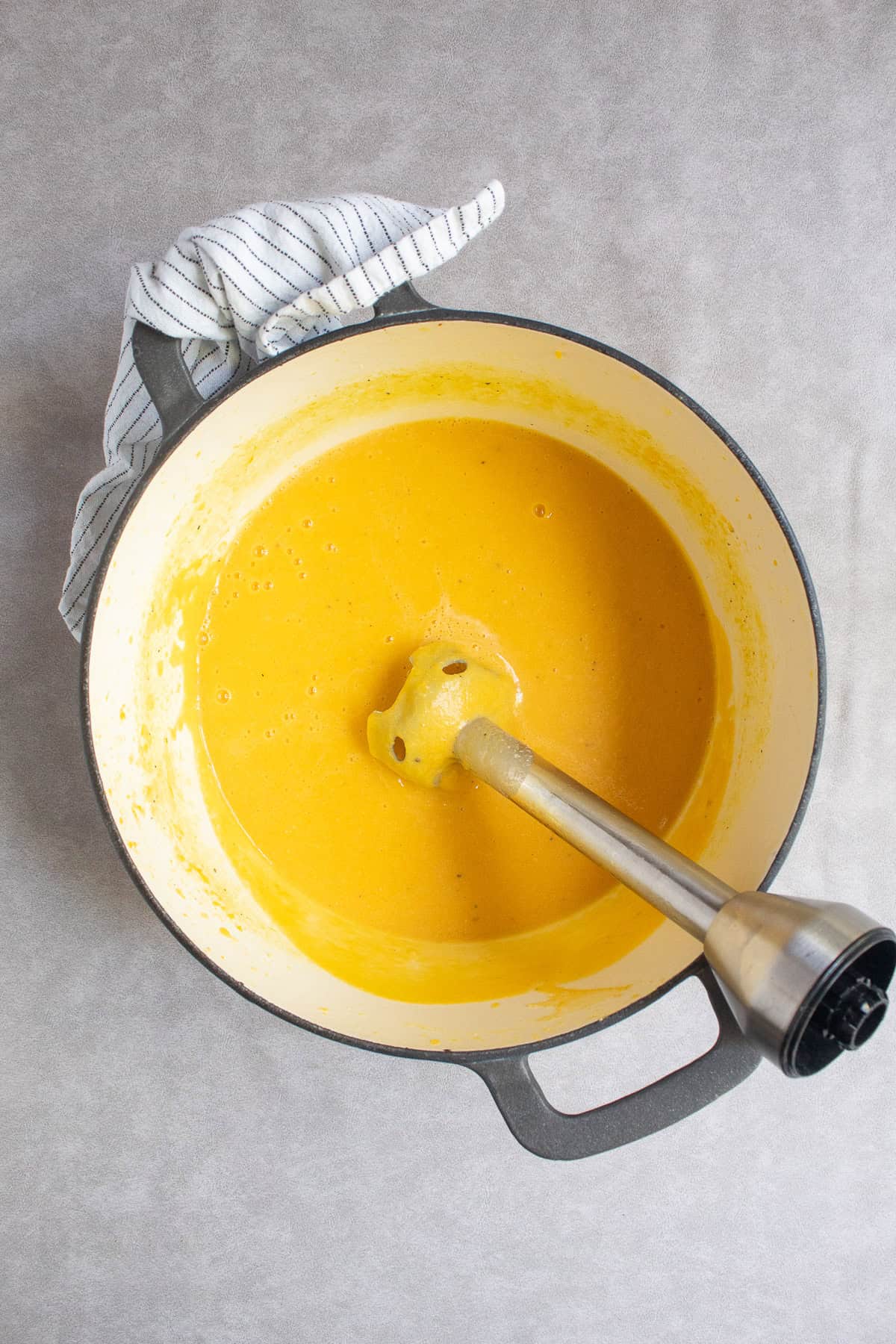 An immersion blender stick sits in the bottom of a pot with a golden yellow sauce.