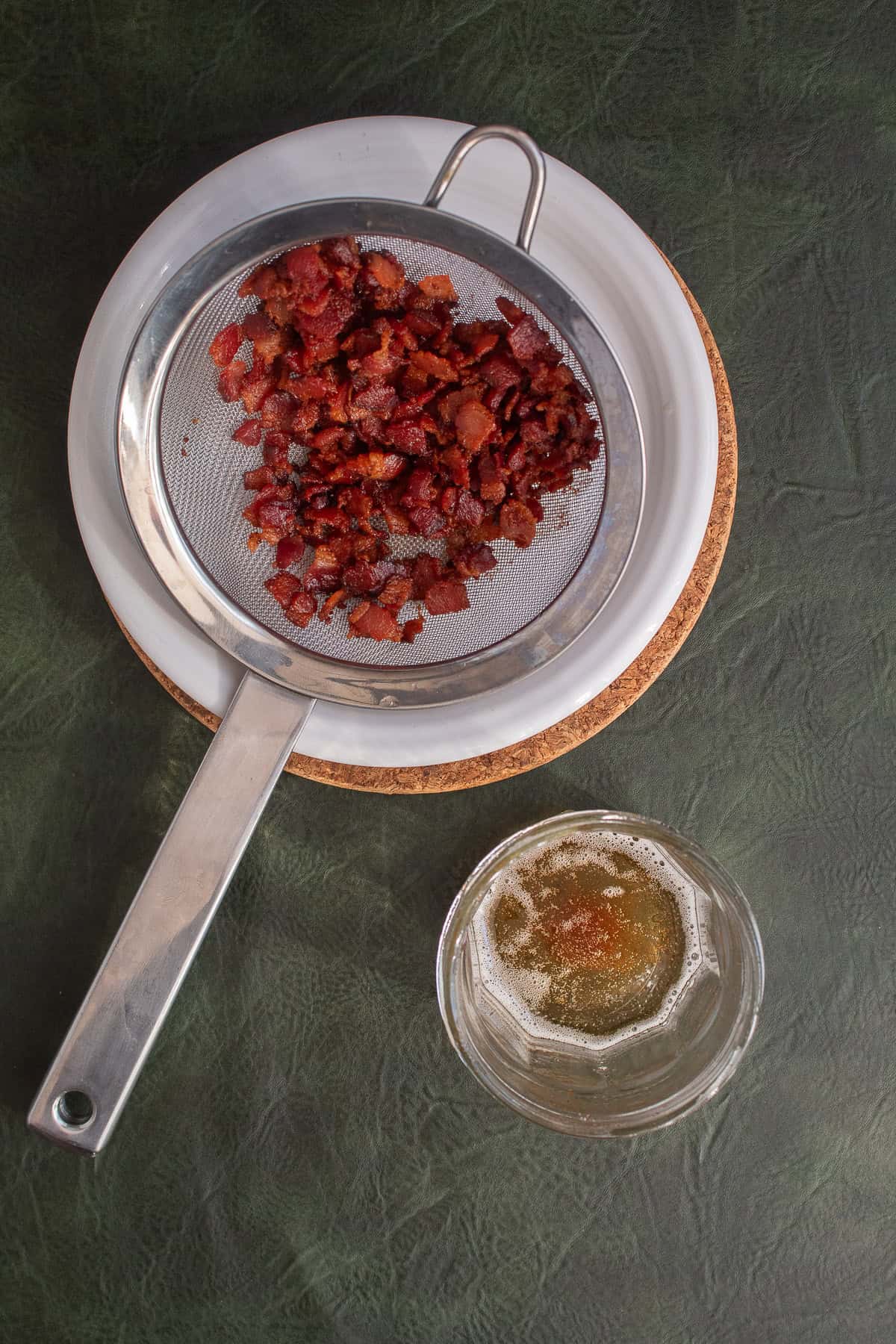 Bacon pieces are drained and the bacon grease is captured in a glass jar.