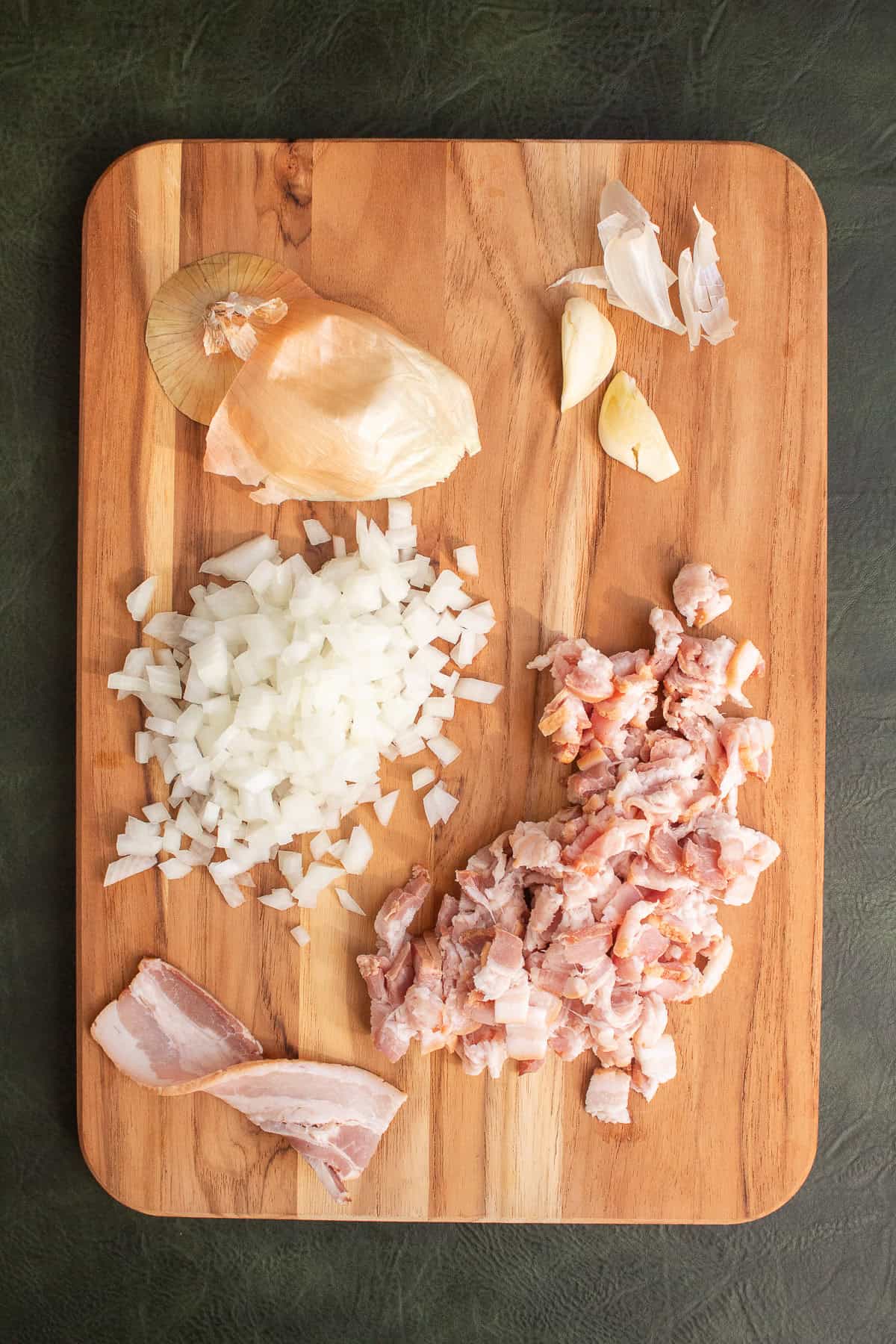 Bacon, onion, and garlic are prepared on a wooden cutting board.