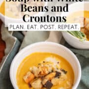 A white bowl of bright orange soup sits on a green striped cloth. A white text box at the top if the image contains the words, "pumpkin soup with white beans and croutons" and "pln. eat. post. repeat."