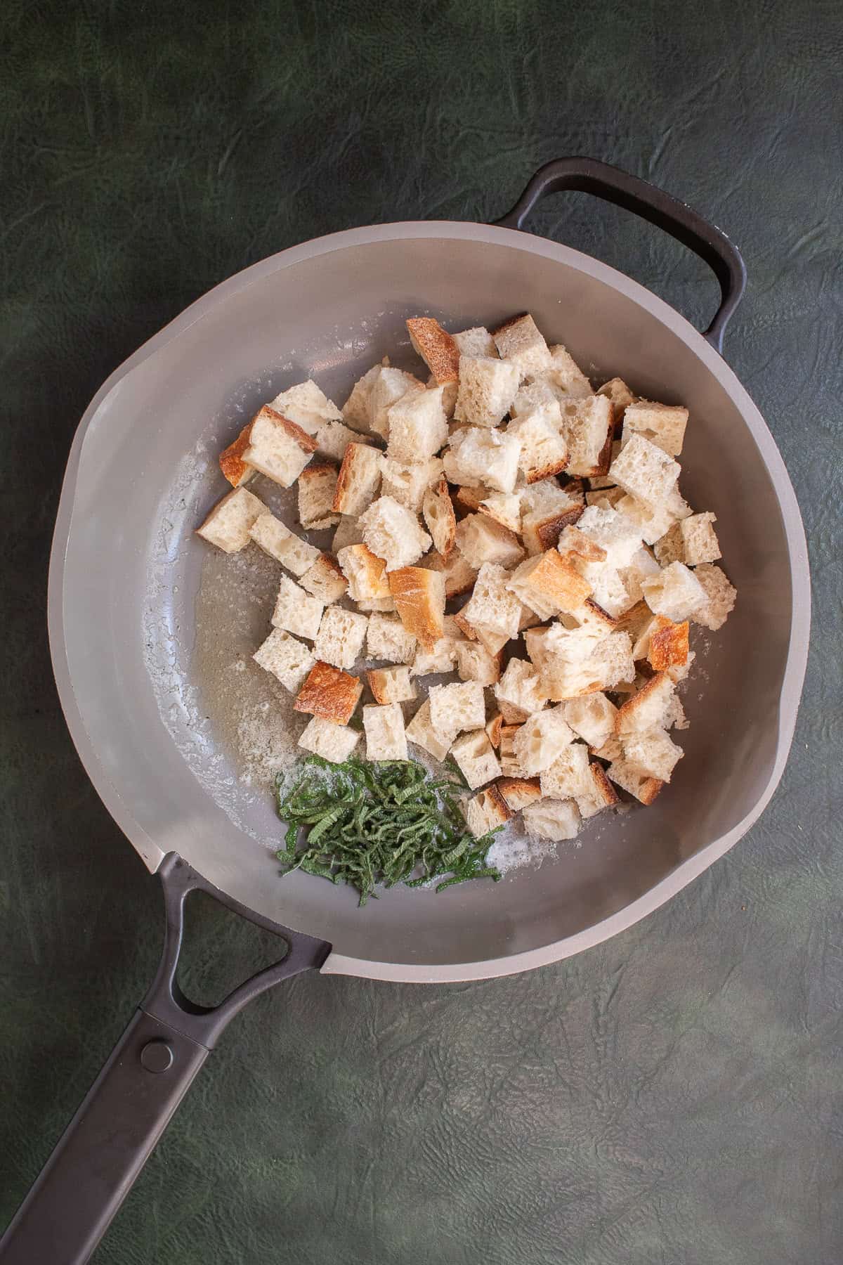 Cubes of bread and sliced sage are added to a light colored skillet.
