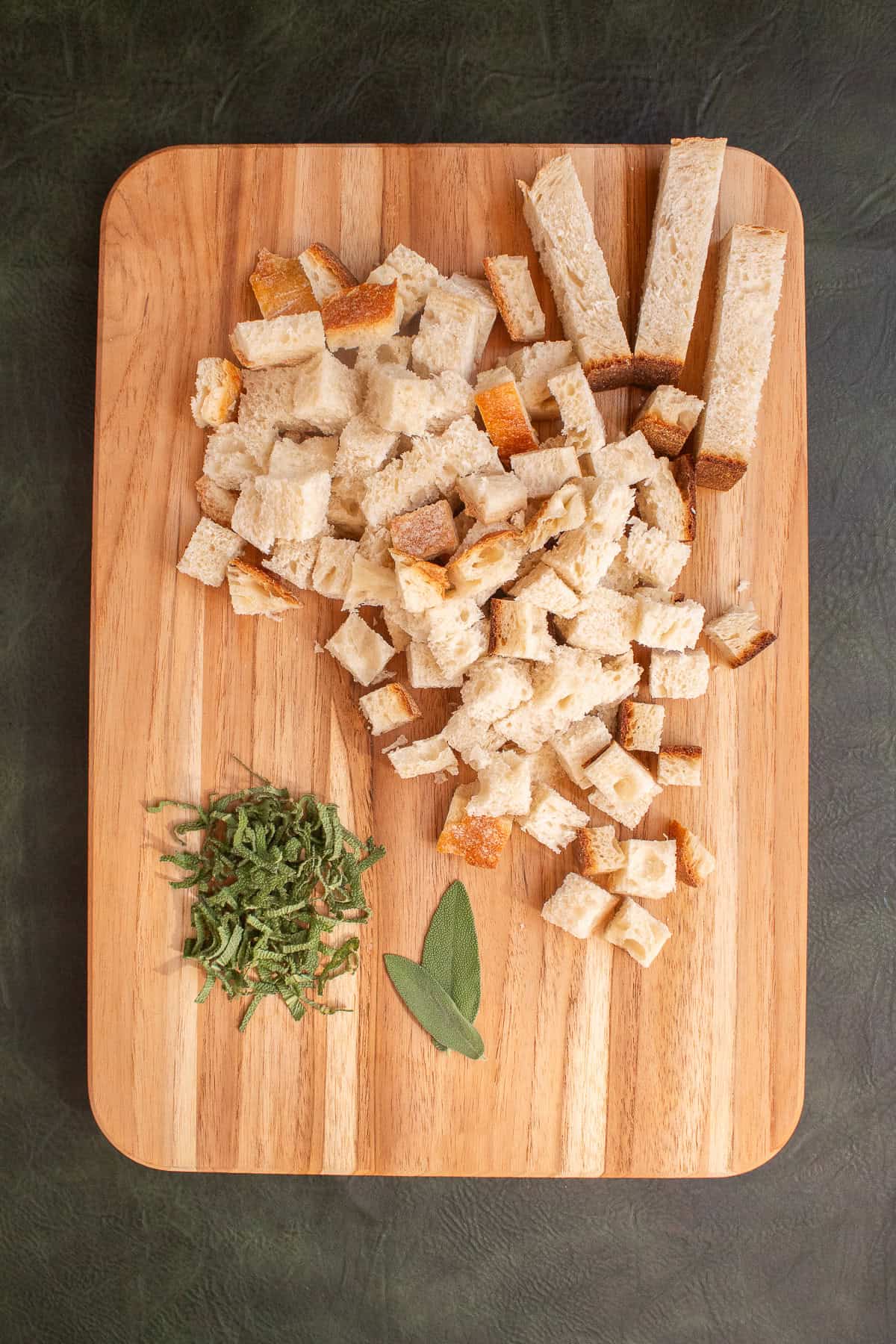 Bread is cut into strips and then cubes next to sliced sage on a wooden cutting board.