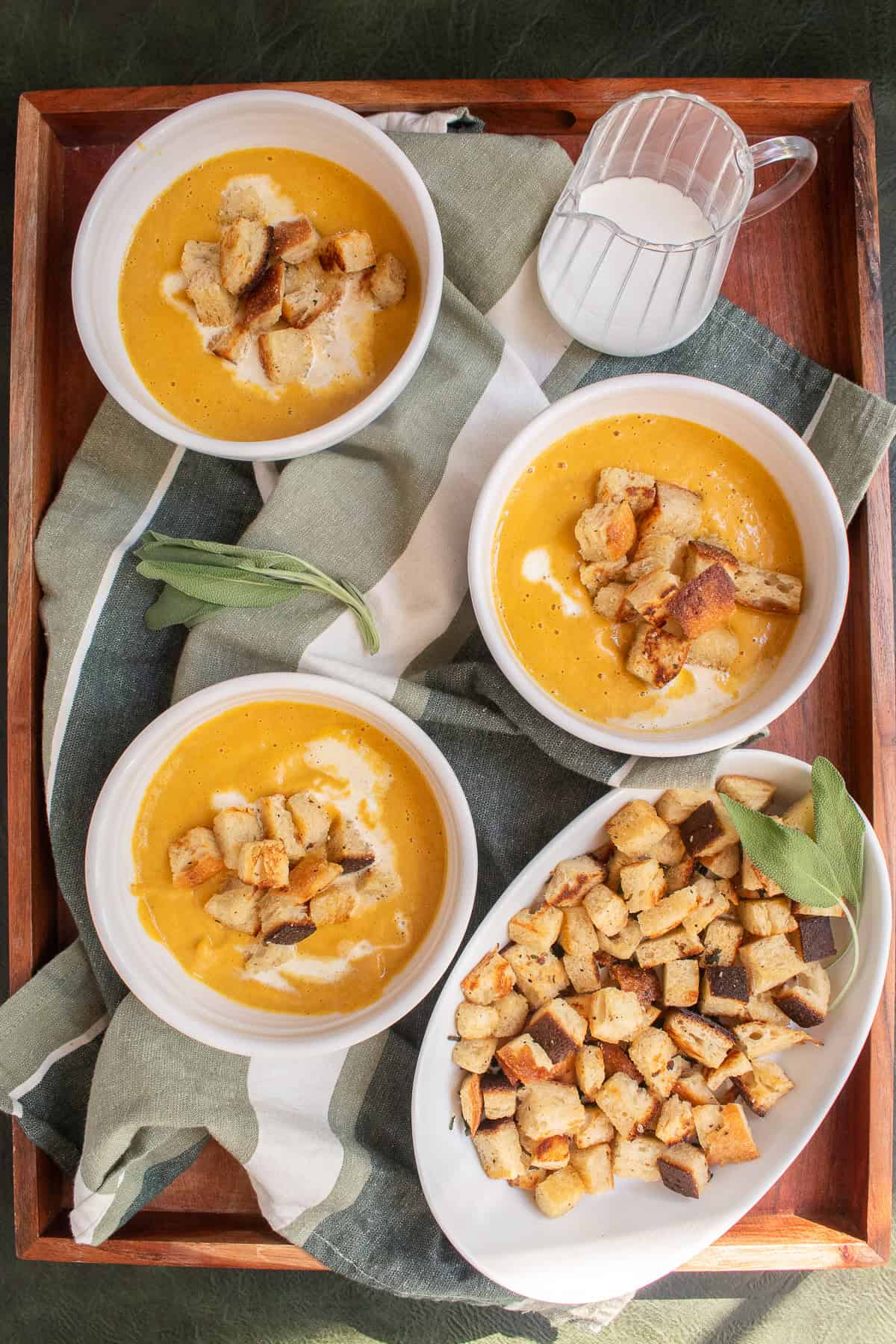 Three bowls of soup sit on a wooden tray alongside a platter of croutons and a small pitcher of half and half.