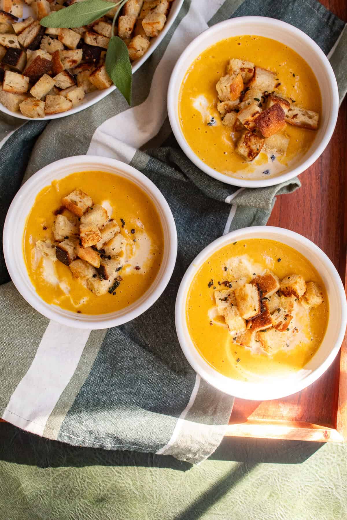Three bowls of bright orange soup are topped with croutons and fried sage.